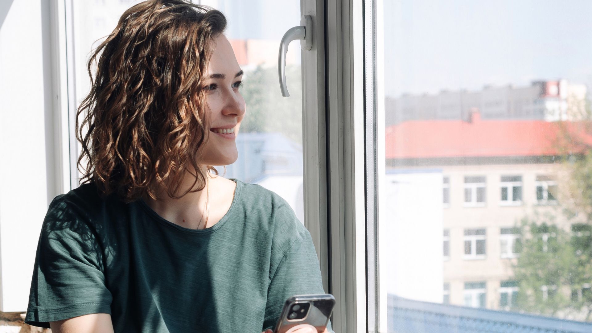 Woman with curly hair looking out a window, holding a phone, with buildings visible outside.