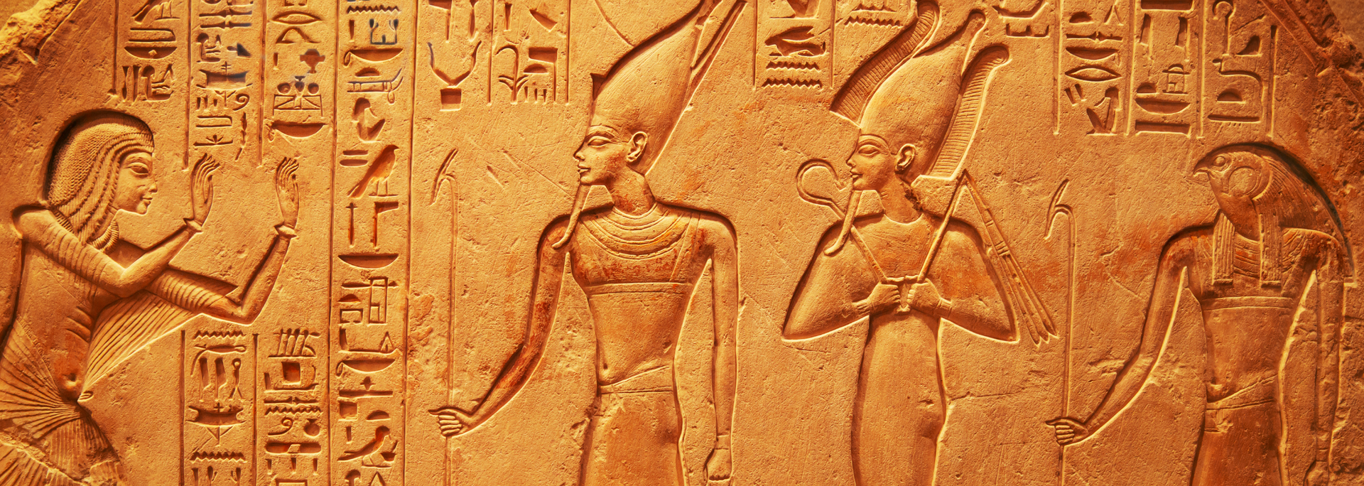 Ancient Egyptian-style carved relief of three figures with hieroglyphs on a tan stone wall