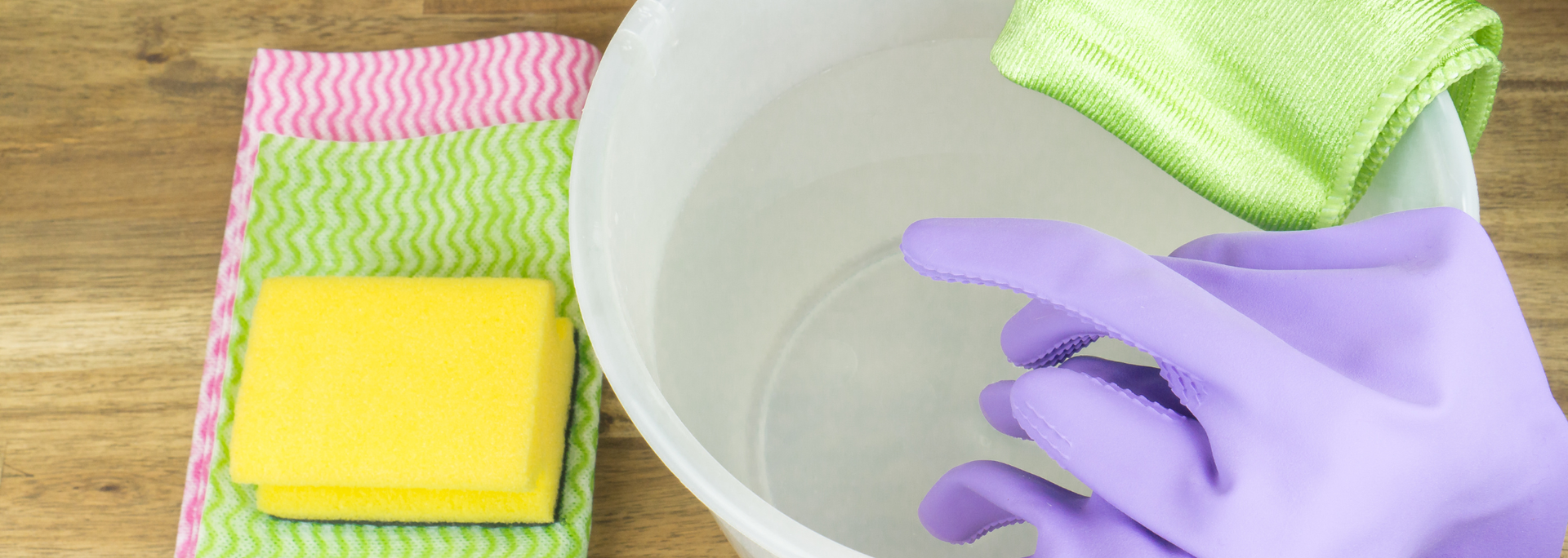 Gloved hand washing dishes in a bowl beside a sponge and colorful cleaning cloths