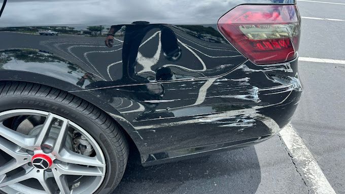 A black car with a damaged bumper is parked in a parking lot.