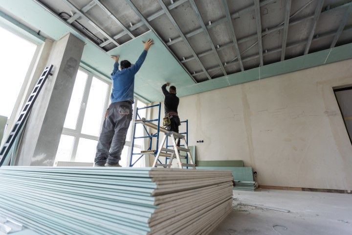 Two workers installing drywall on a ceiling in a building under construction.