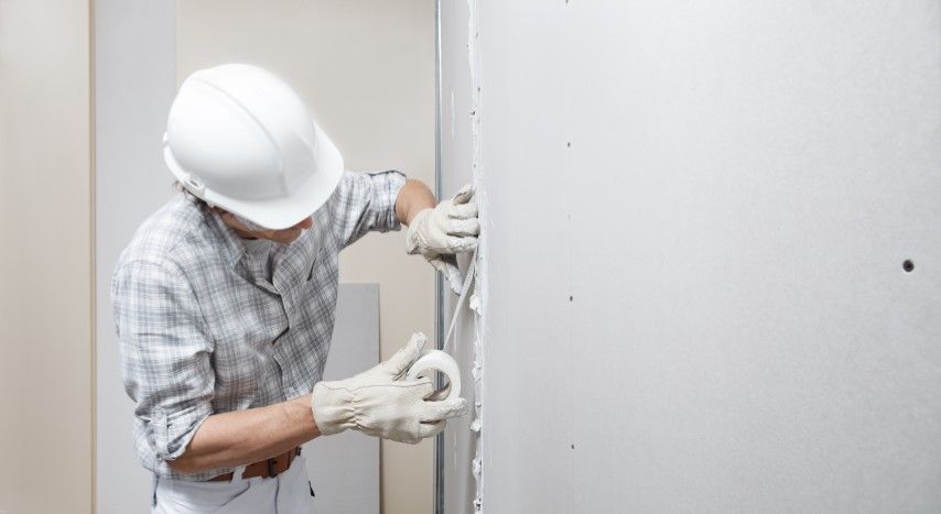 Person in a white hard hat and gloves applying tape to drywall in a room.
