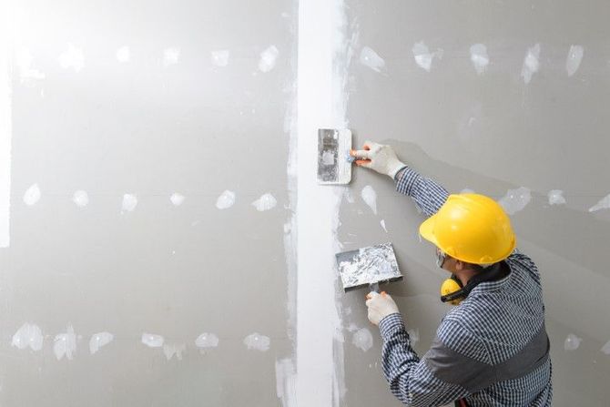 Worker in a yellow hard hat applying joint compound to drywall with a trowel, in a room.