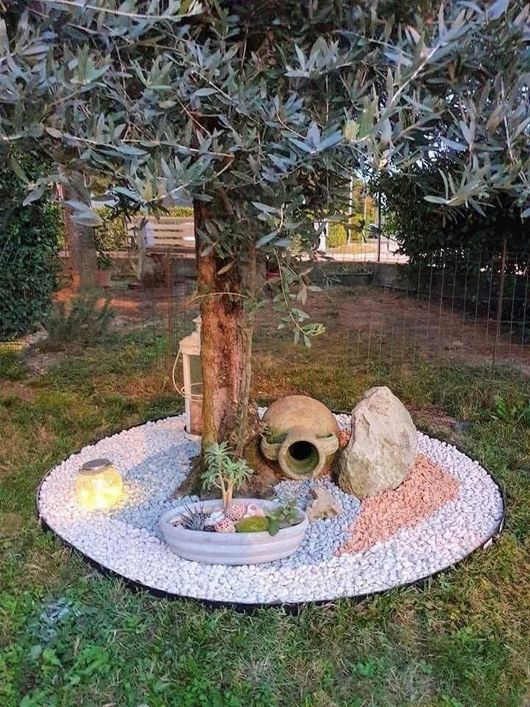 Un tronco d'albero circondato da pietre bianche, su cui poggiano una lanterna, una piccola pianta in vaso, una brocca di terracotta e una grande roccia.