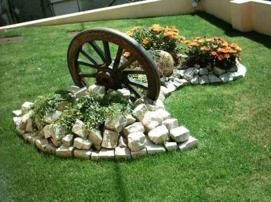 Una ruota di carro in legno si erge come elemento decorativo del giardino, circondata da pietre, vegetazione lussureggiante e fiori arancioni in fiore.