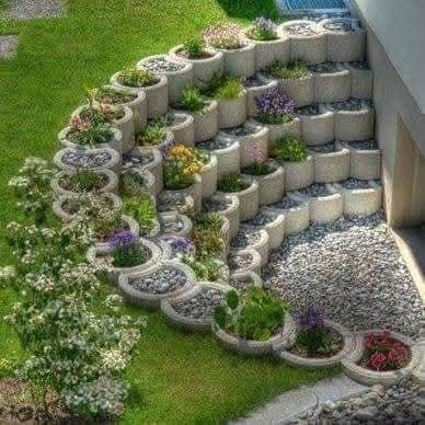Un muro di contenimento curvo a più livelli, realizzato con fioriere in cemento riempite con vari fiori e arbusti, adiacente al muro di una casa.
