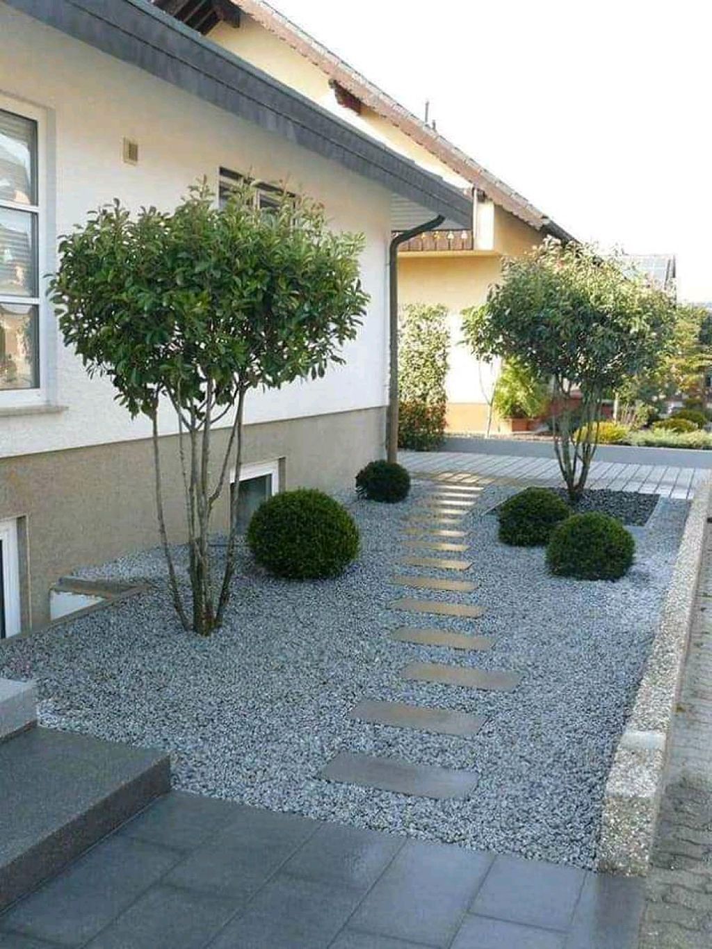 Un sentiero di ghiaia costeggia una casa bianca, con piccoli arbusti rotondi e due alberi ornamentali sotto un cielo luminoso.