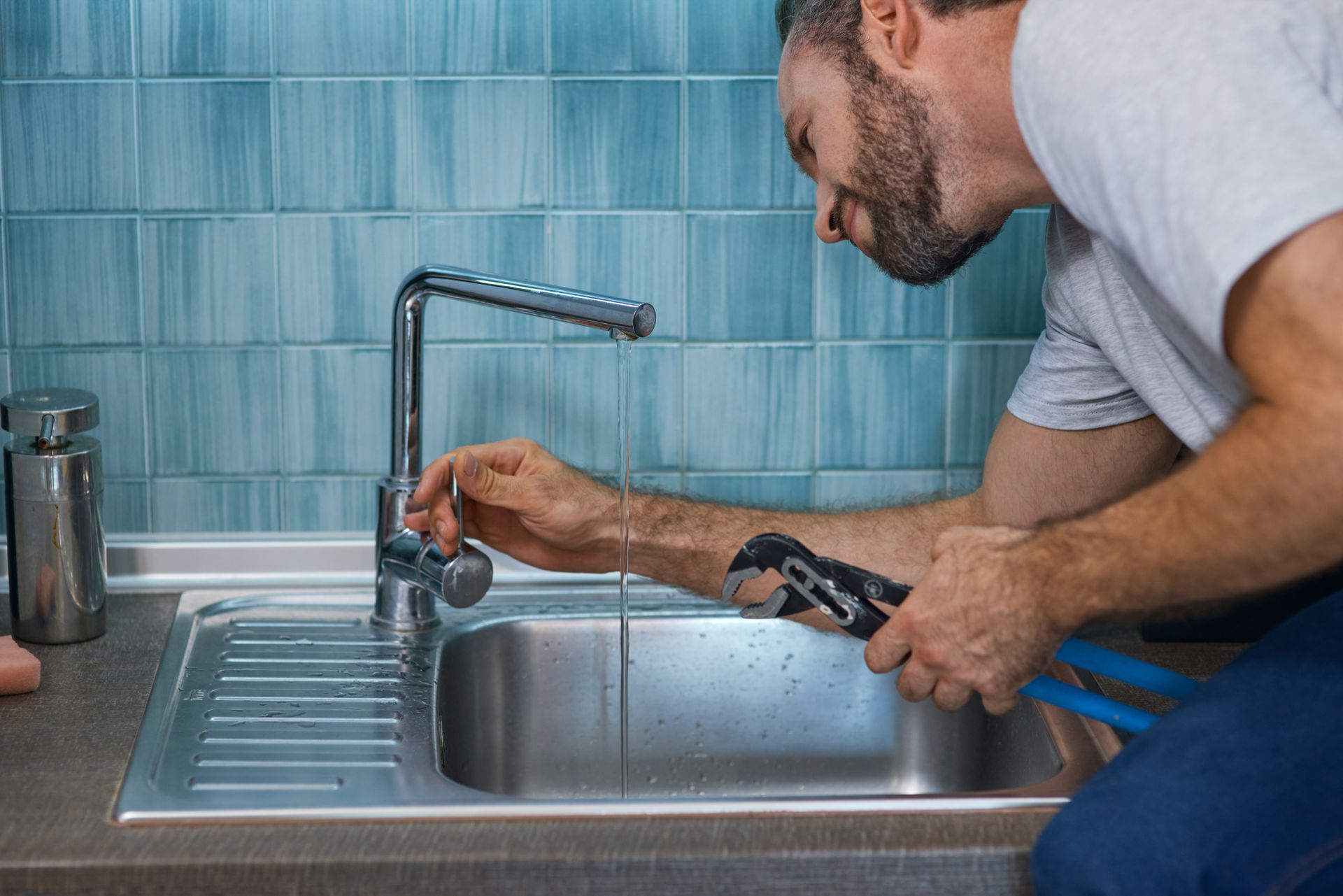 Plumber repairing a kitchen sink faucet with a wrench beside a blue-tiled backsplash