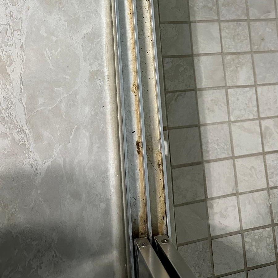 Close-up of a dirty metal shower door track beside white wall and tiled floor