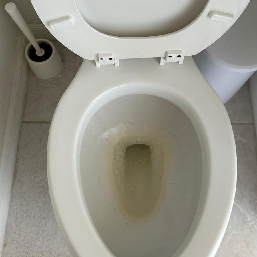 Open toilet bowl with light staining in a small bathroom, toilet brush visible beside it