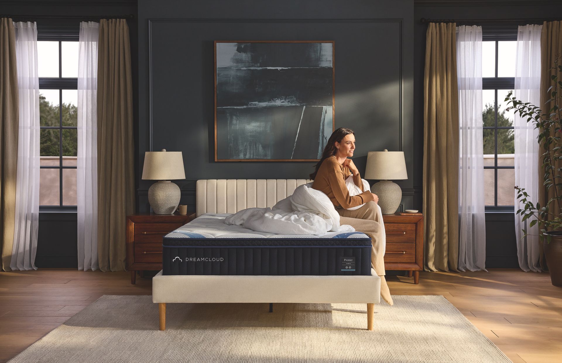 Woman sitting on a bed in a dark bedroom, surrounded by neutral-colored decor; mattress on the bed.
