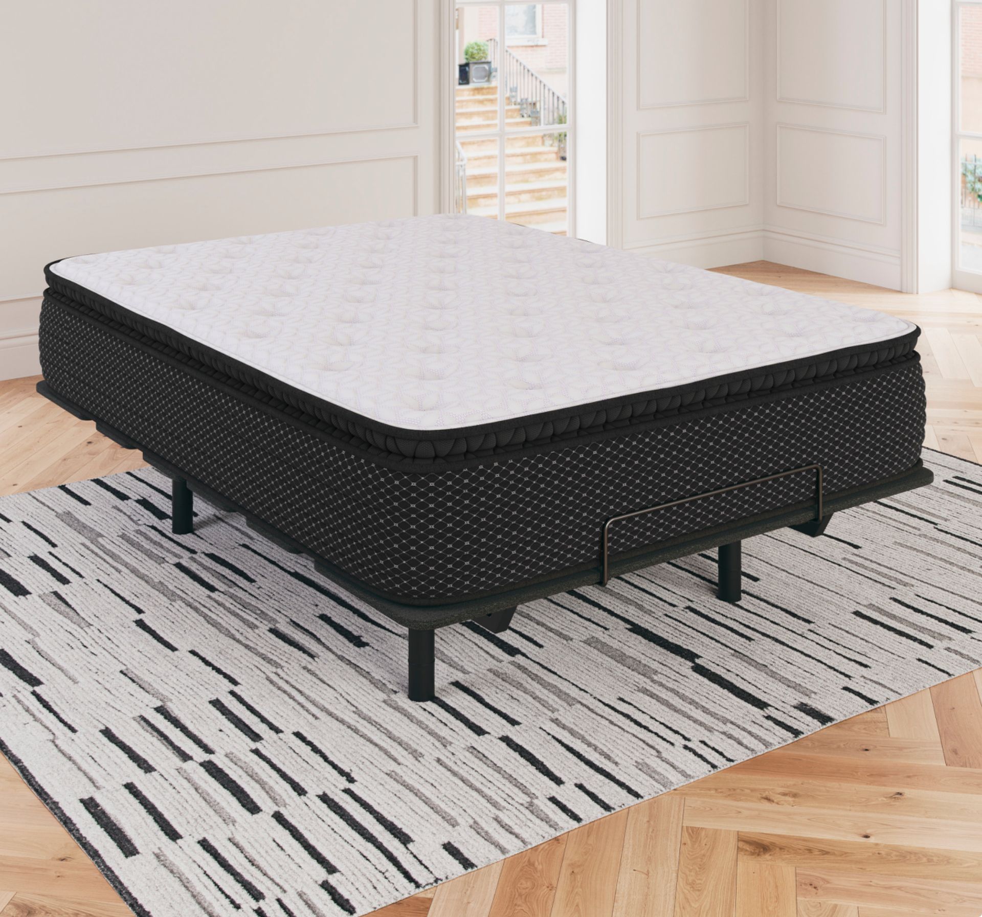 Black and white mattress on a metal frame, set on a patterned rug in a bright room.