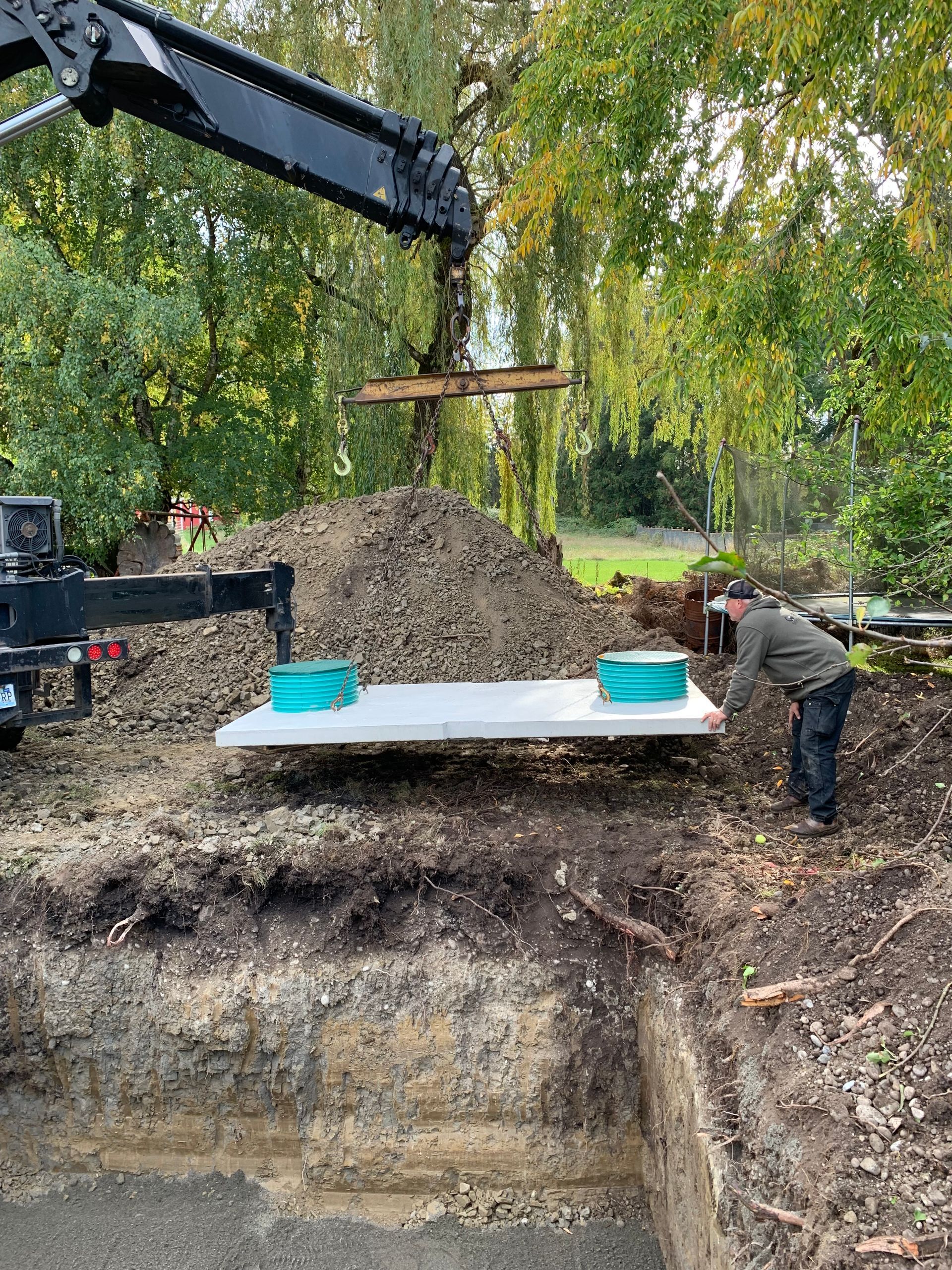 Man in trench with pipes, excavator, and mountain in background.