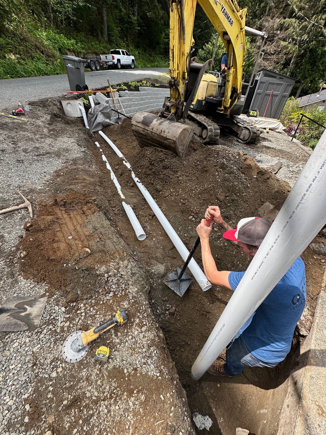 Man in trench with pipes, excavator, and mountain in background.