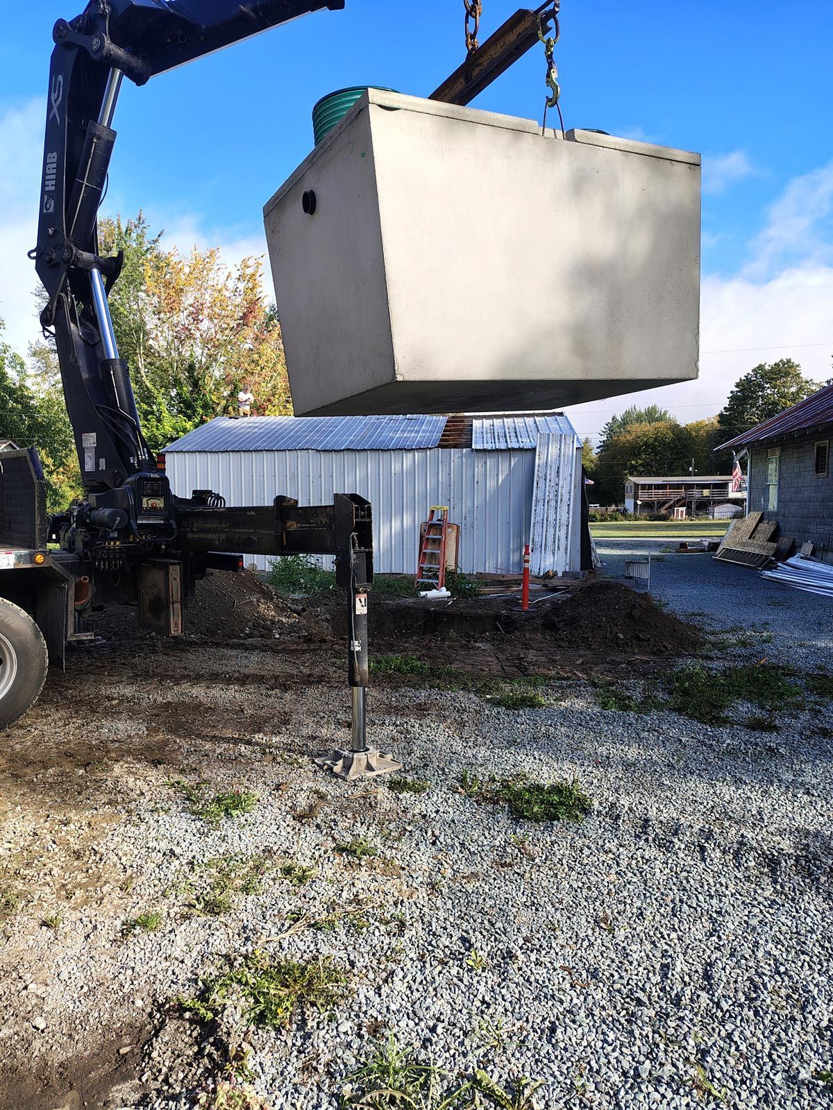 A crane lifting a large concrete septic tank in a gravel yard near a shed and trees.