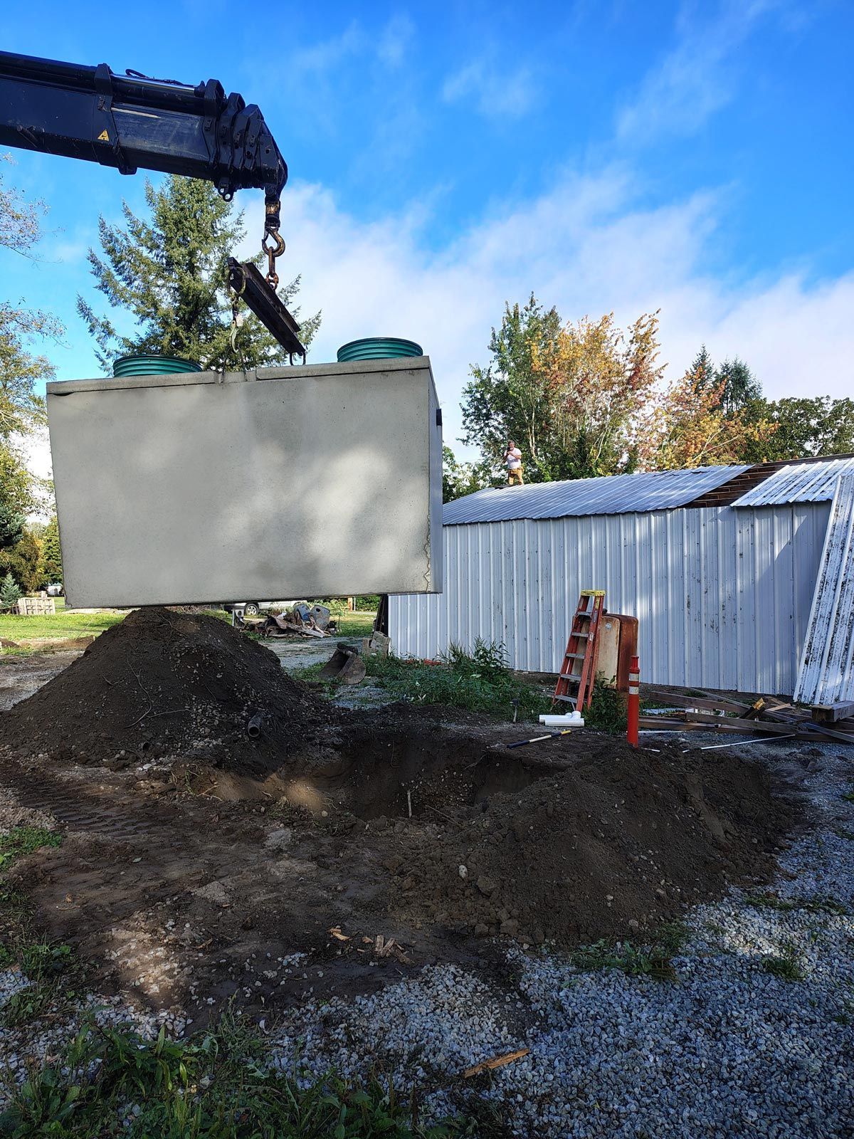 Crane lifting a large concrete septic tank over a dirt mound. The sky is blue.
