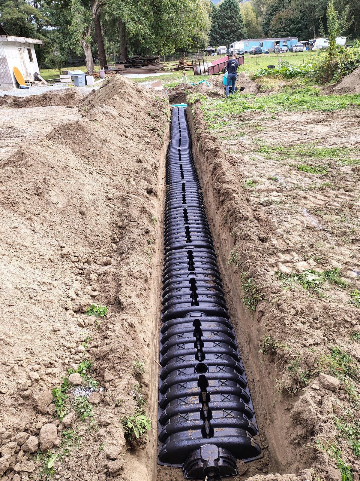 A black drainage system installed in a trench, surrounded by dirt and grass, with a worker in the background.