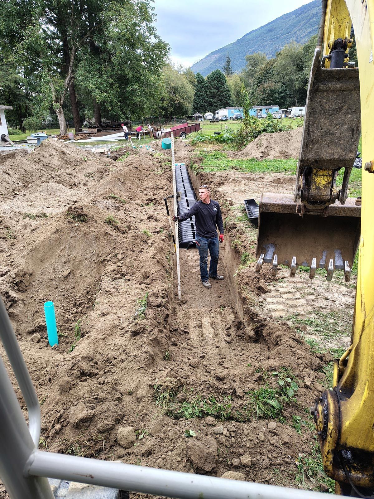Man in trench with pipes, excavator, and mountain in background.
