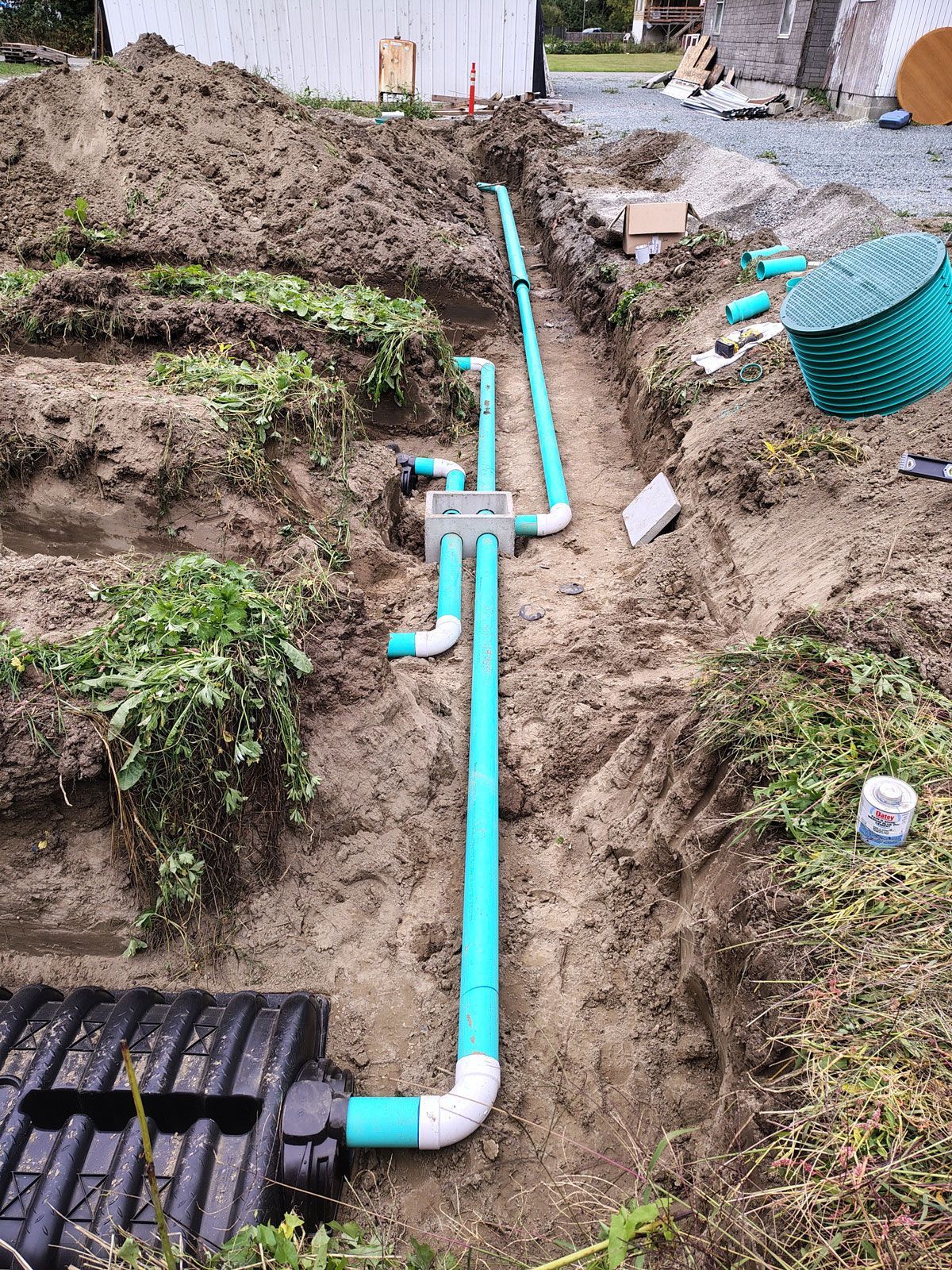 Plumbing installation in a trench: turquoise pipes, a black tank, and a green lid are being installed outdoors.