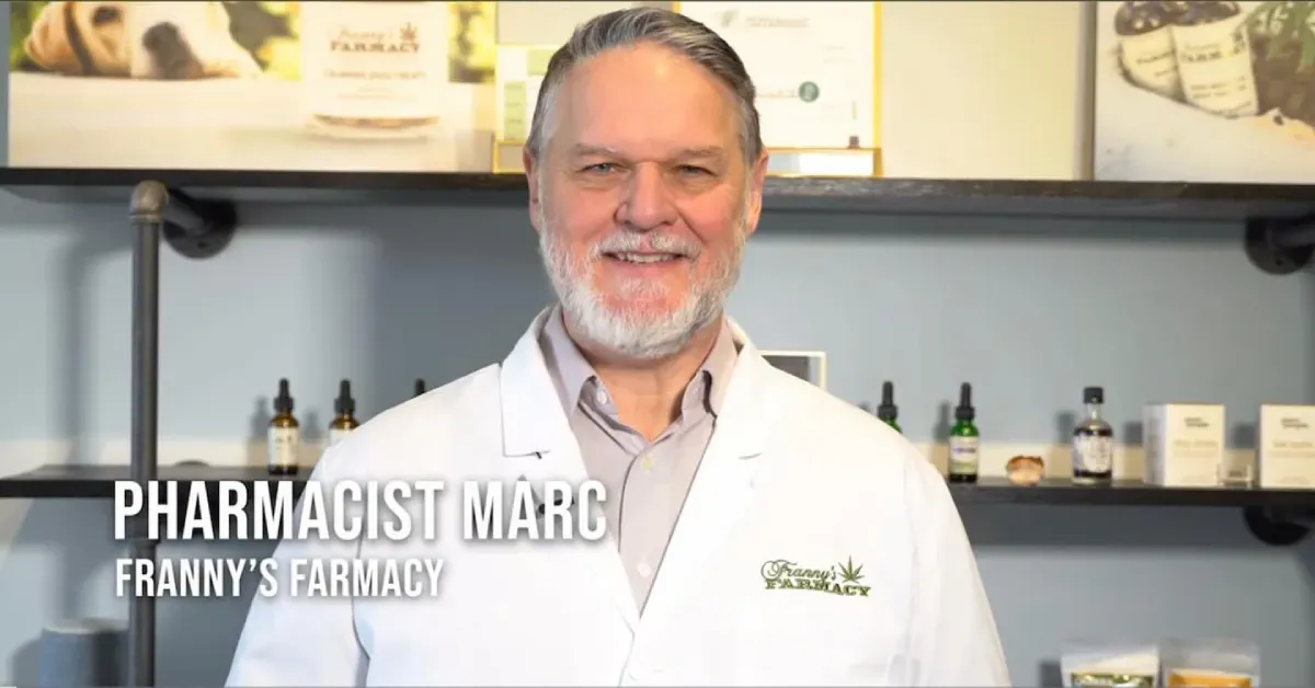 Pharmacist Marc in a white coat at Franny's Pharmacy, smiling in front of products on shelves.