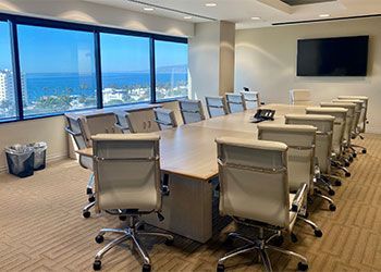 An office conference room with a long table and chairs, overlooking an ocean view through large windows. A TV is mounted on the wall.
