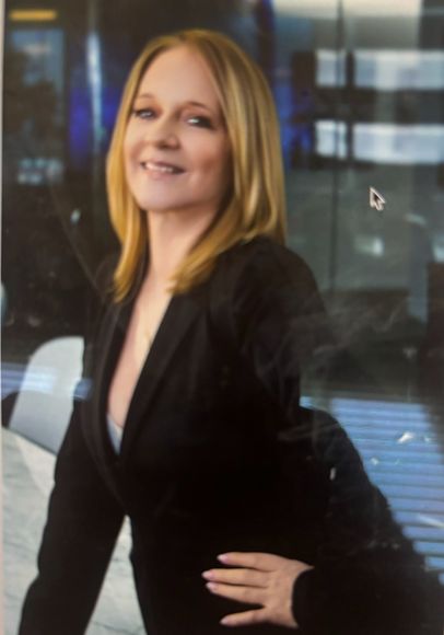 Woman with blonde hair, smiling, wearing a black blazer, leaning against a glass wall.