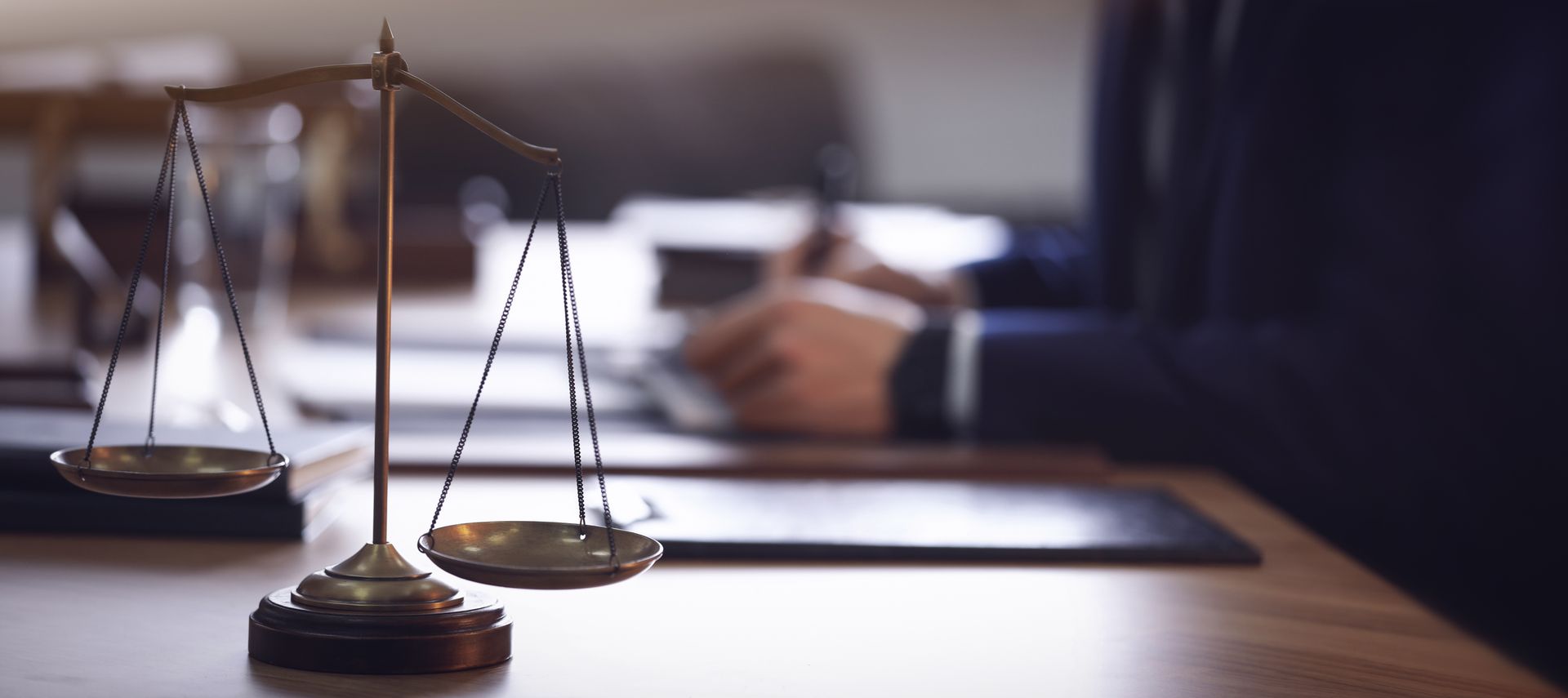 A justice scale on a wooden desk with a blurred person wearing a navy suit.