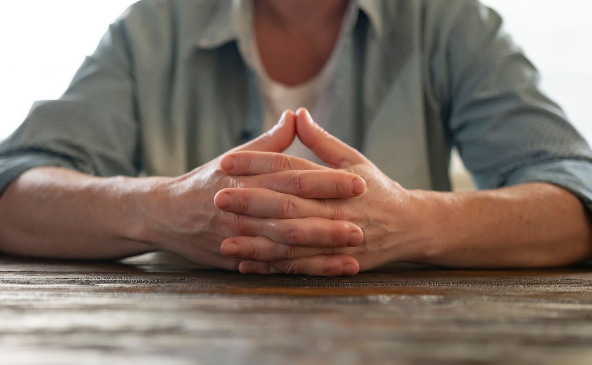 Person's hands clasped on a wooden table, palms up, fingers touching, wearing a blue shirt.