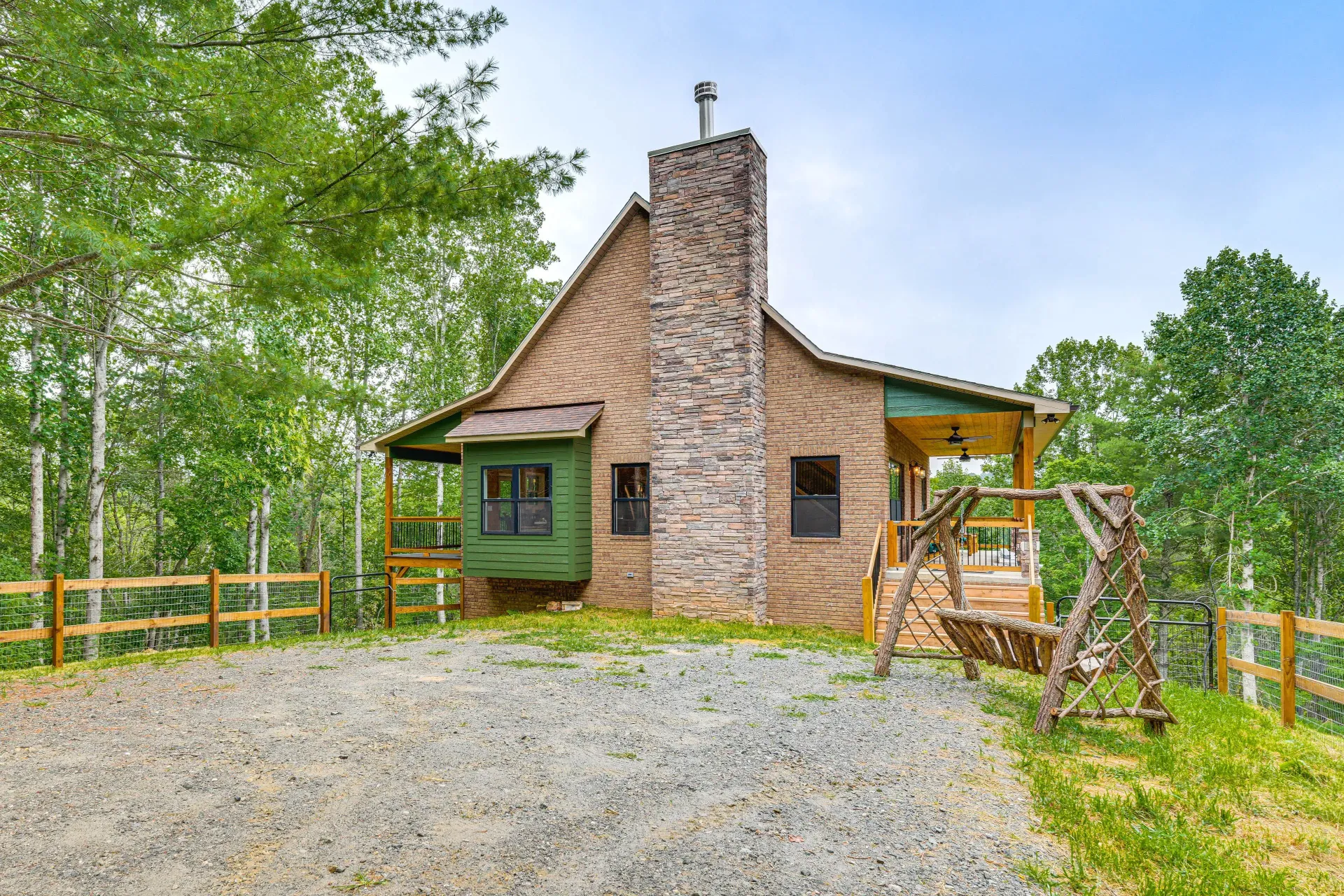 Rustic cabin with stone chimney and green trim beside a gravel drive, surrounded by trees and wooden fencing