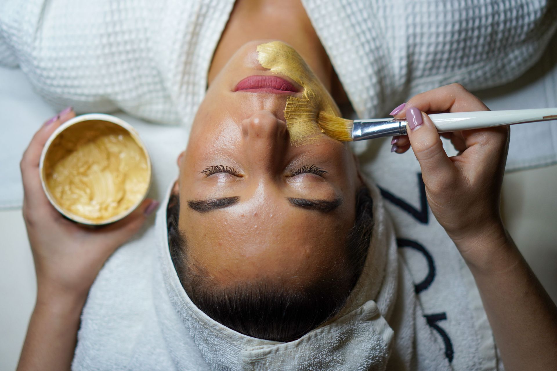 Facial mask applied with a brush to a reclining person at a spa, with a cream bowl nearby
