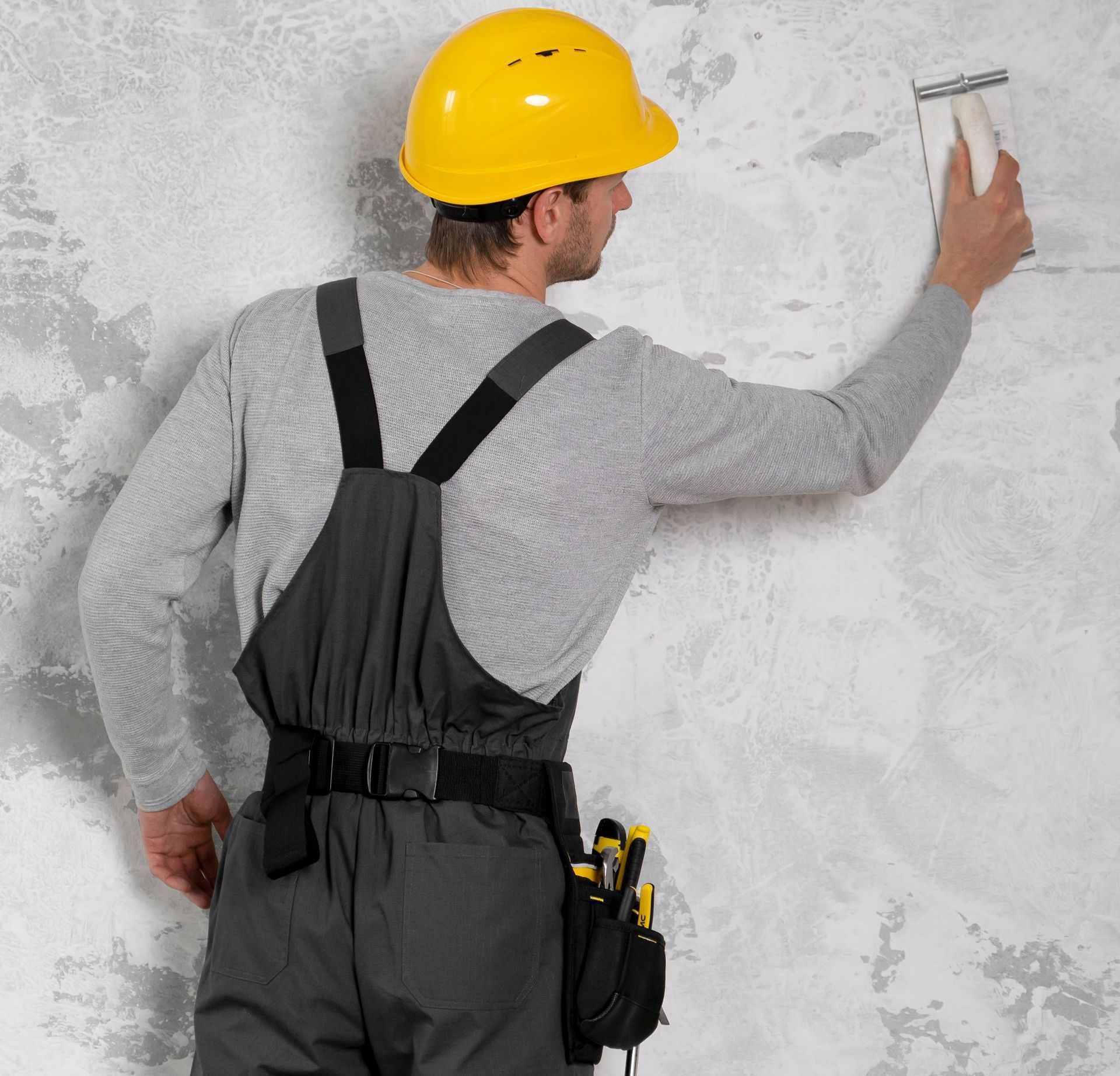 a man wearing a yellow hard hat and overalls is holding a tool against a wall