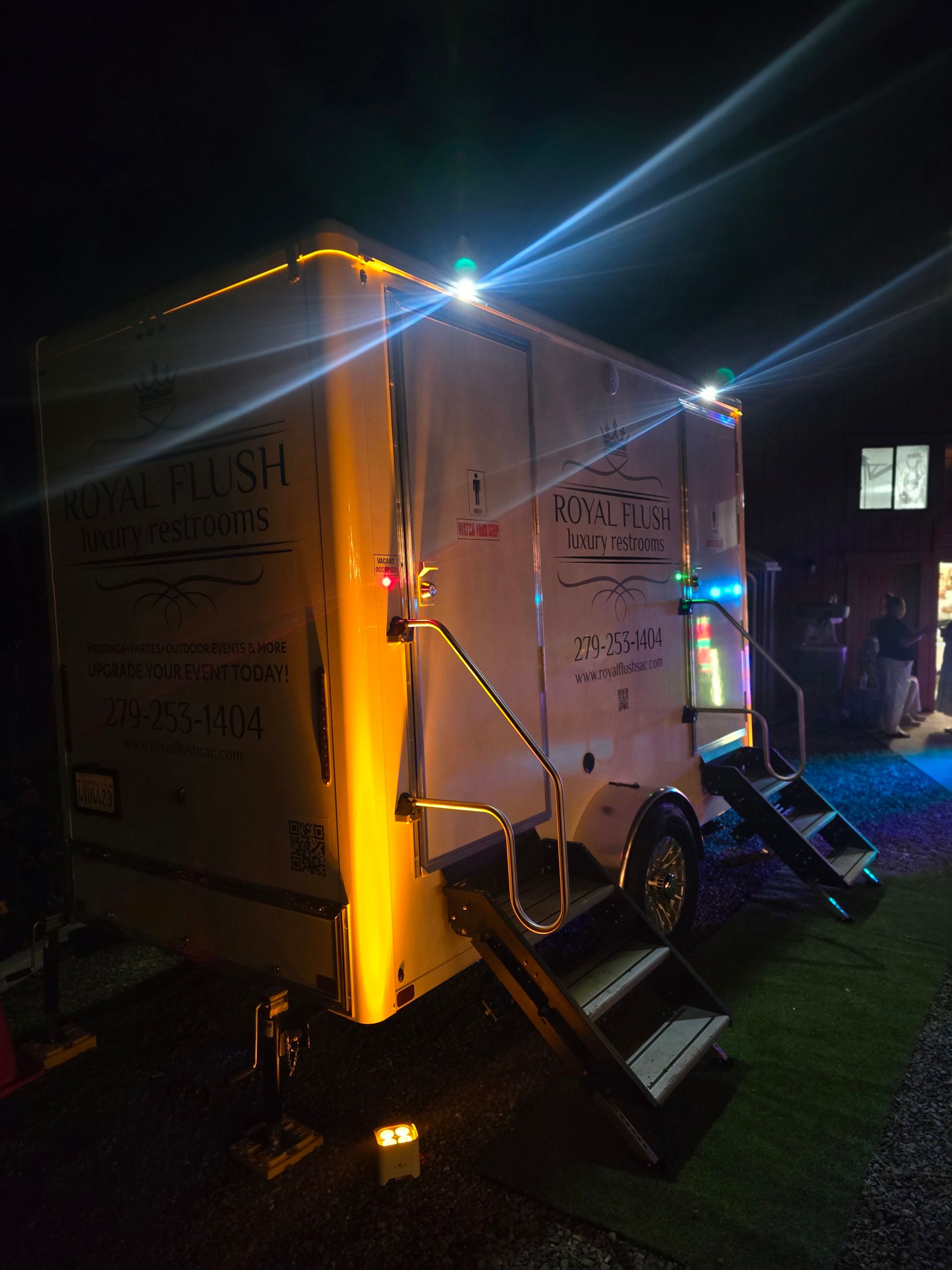 Nighttime view of a lit white trailer with steps and chain barriers, parked on grass at an outdoor event.