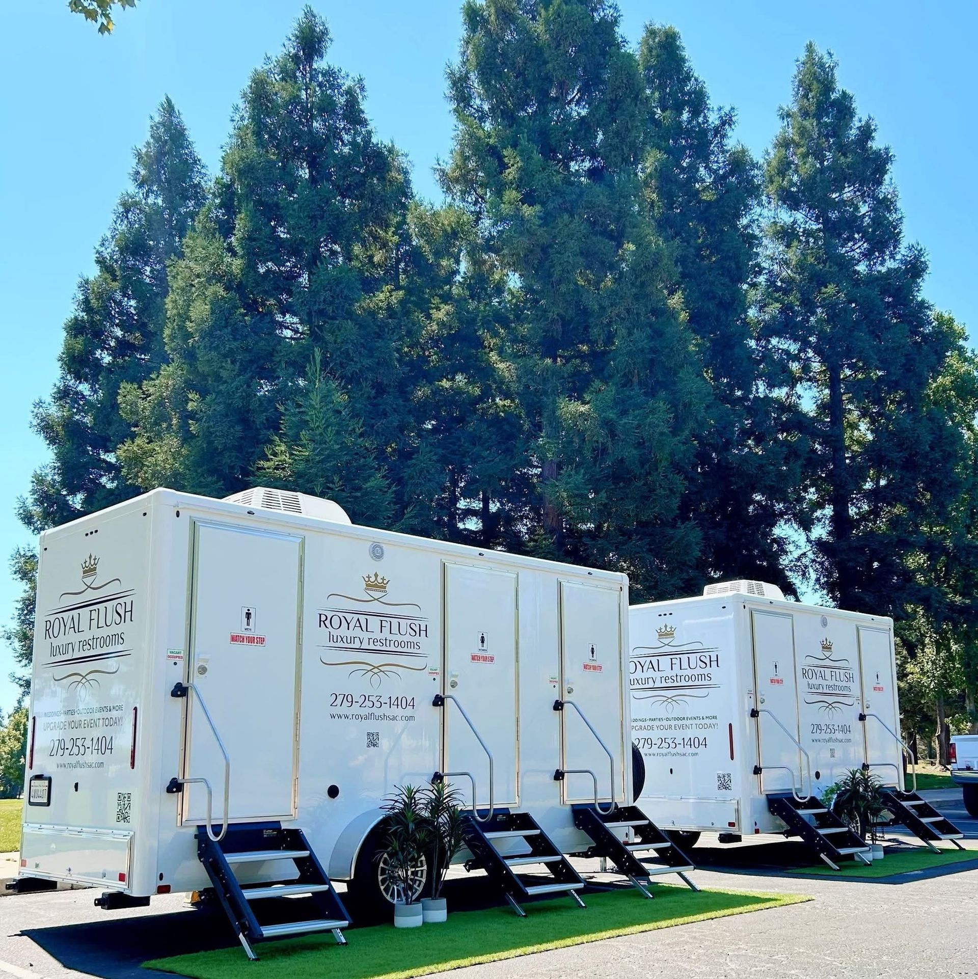White portable restroom trailers on grass beneath tall trees outdoors