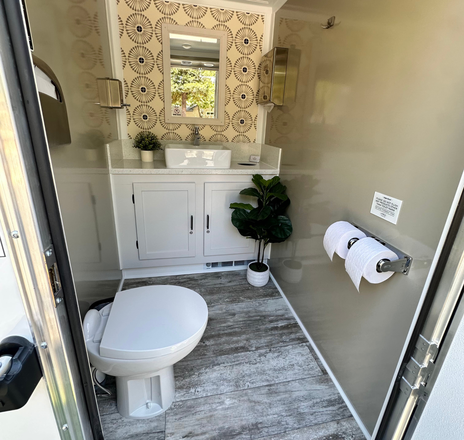 Luxurious portable toilet with white vanity, patterned wallpaper, and faux plant.