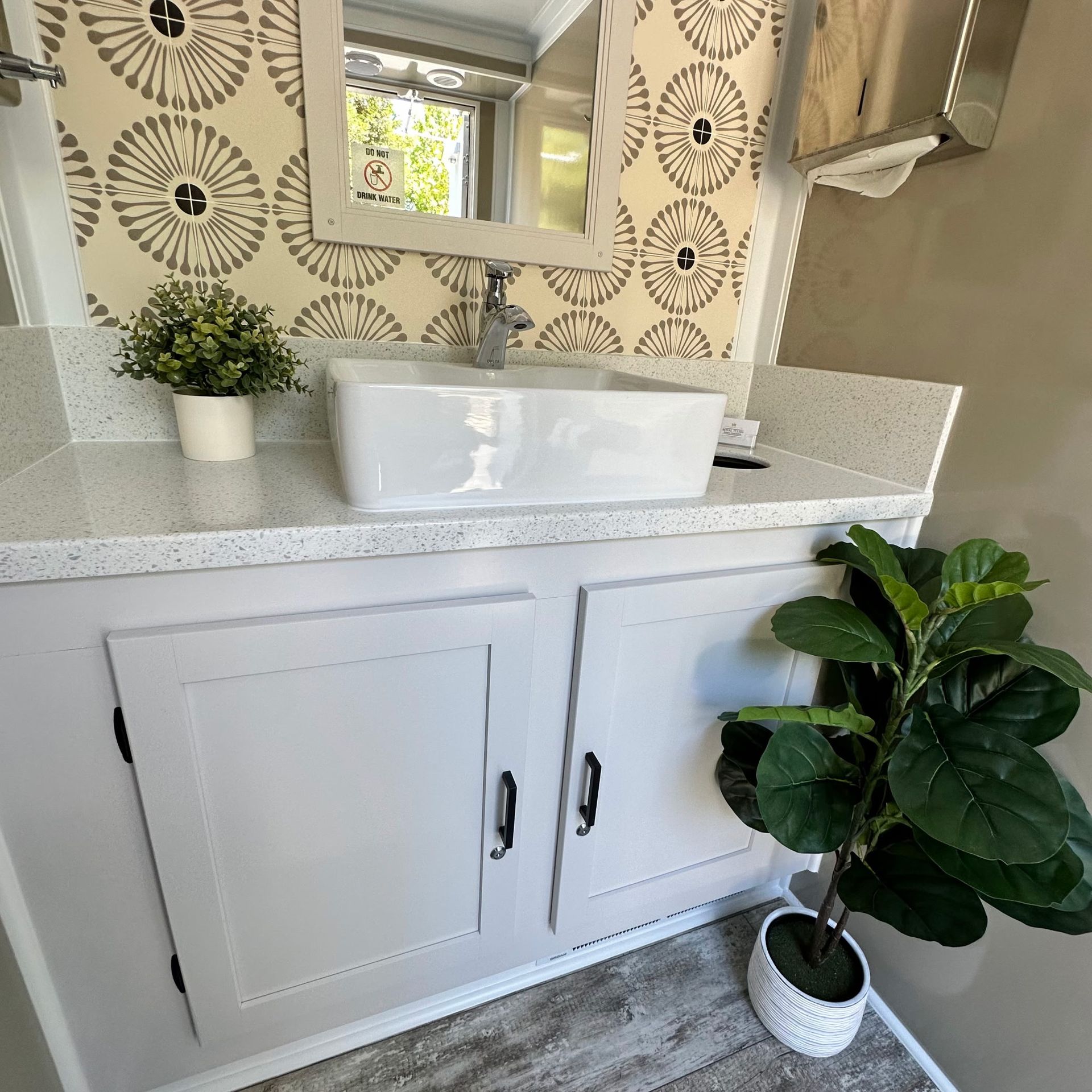 White bathroom vanity with vessel sink, patterned wallpaper, mirror, and potted plants.
