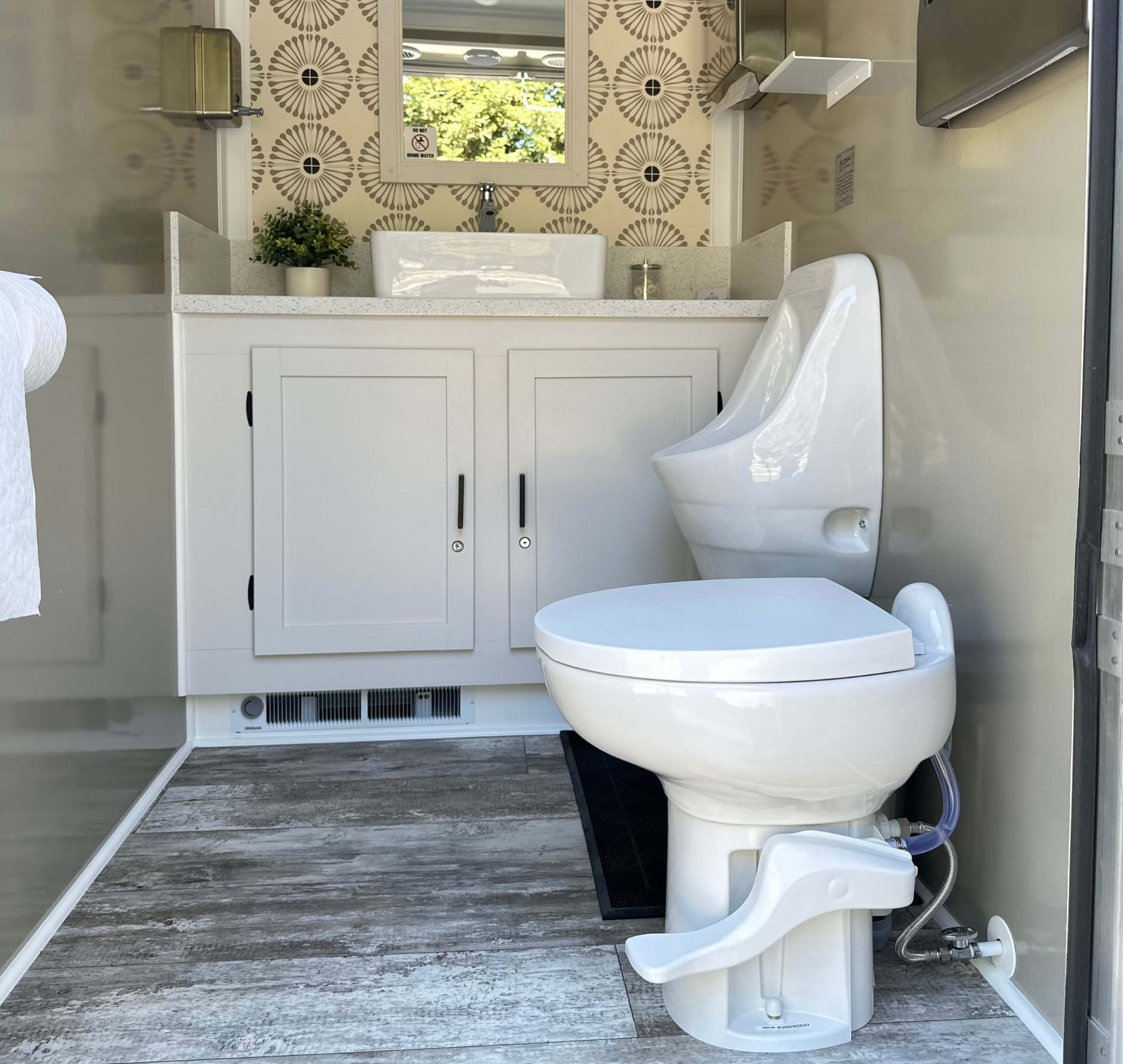 Interior of a modern white portable toilet with a sink, mirror, and urinal. Grey wood-look flooring.