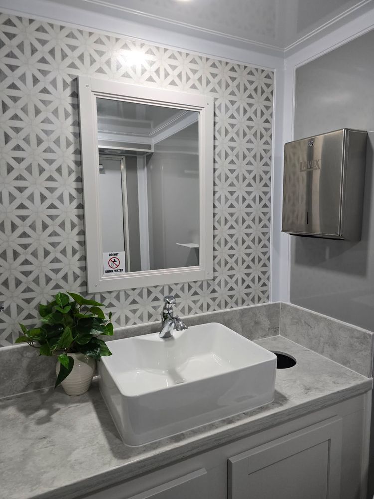 A modern bathroom with a sink, mirror, and paper towel dispenser; decorative wallpaper and a potted plant.