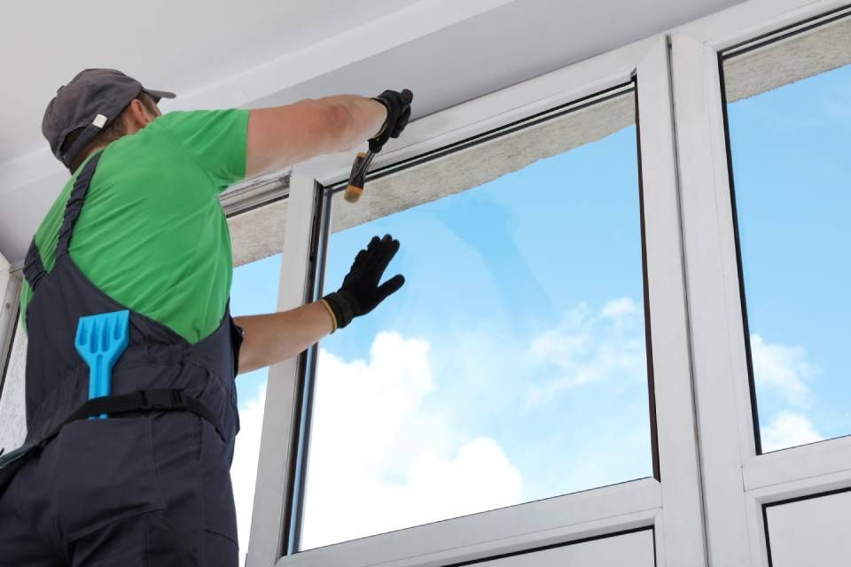 A person in a green shirt and work overalls applies sealant with a tool to the upper frame of a white plastic window.