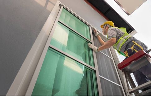 A worker in a yellow safety vest and hard hat installs a large glass window on a building exterior while on a ladder.