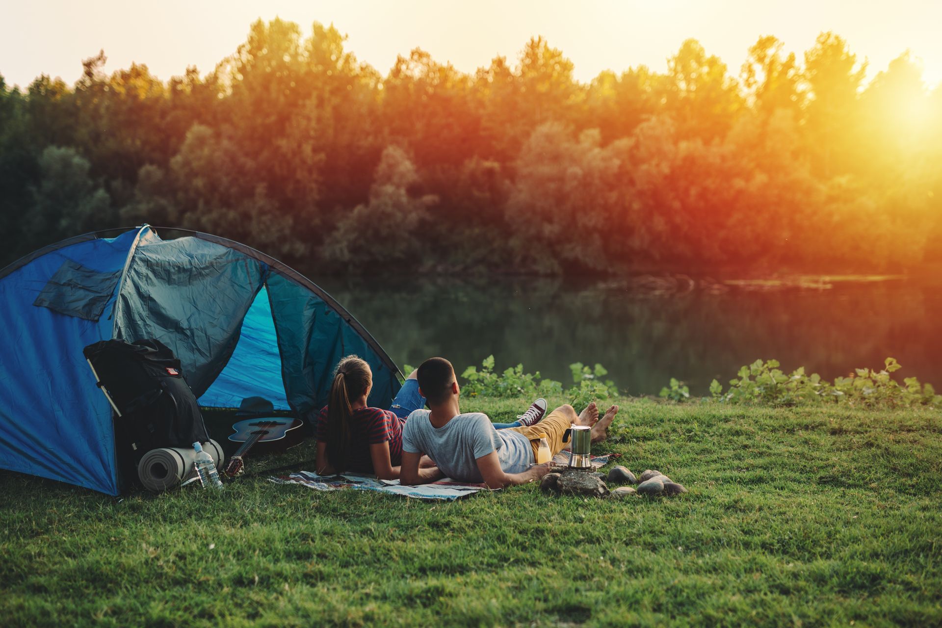 Two campers sitting by a blue tent at sunset beside a lake, with a picnic on the grass