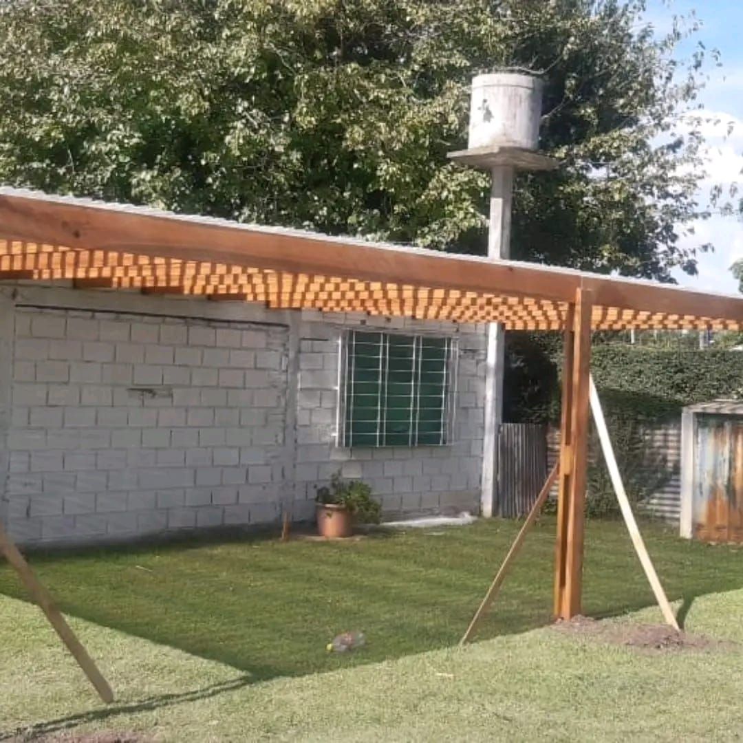 Pérgola de madera sobre un edificio de ladrillo blanco sobre un césped; tanque de agua al fondo.