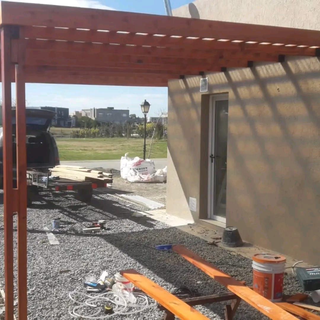 Construcción de pérgola de madera junto a una casa con puerta abierta. Remolque con entrada de grava y madera.