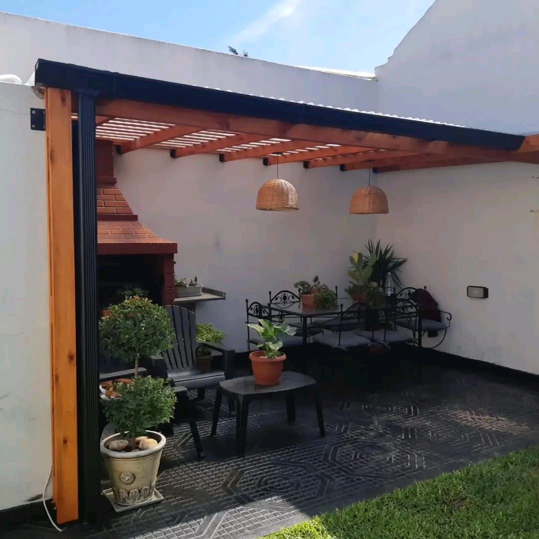 Patio con pérgola de madera, parrilla, asientos, plantas y luces colgantes.