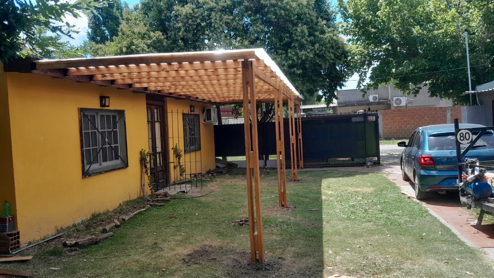 Casa amarilla con pérgola de madera, patio con césped y coche azul aparcado cerca.