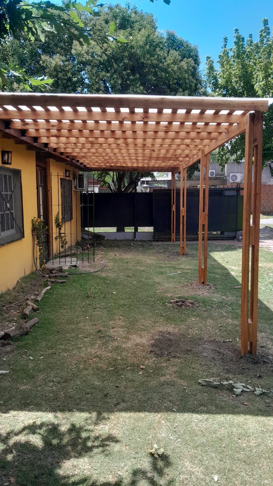 Pérgola de madera que cubre la puerta y la ventana exterior de una casa amarilla, con césped verde.