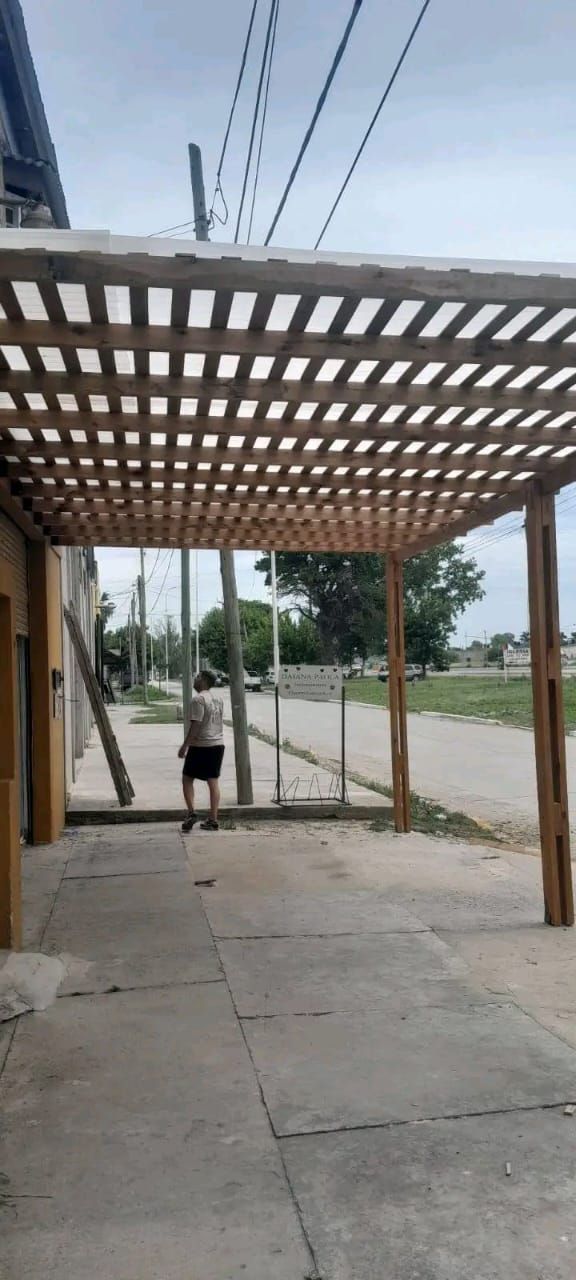 Una persona camina bajo una pérgola de madera con techo de rejilla. Al otro lado, se ve una calle y una puerta. Día nublado.