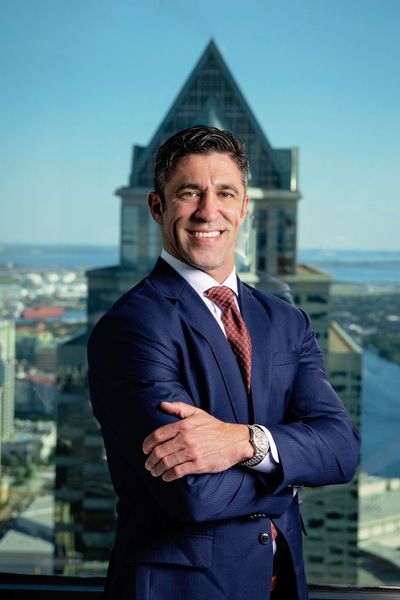A man in a suit and tie is standing with his arms crossed in front of a tall building.