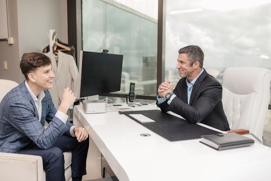 A man is sitting at a desk talking to another man.