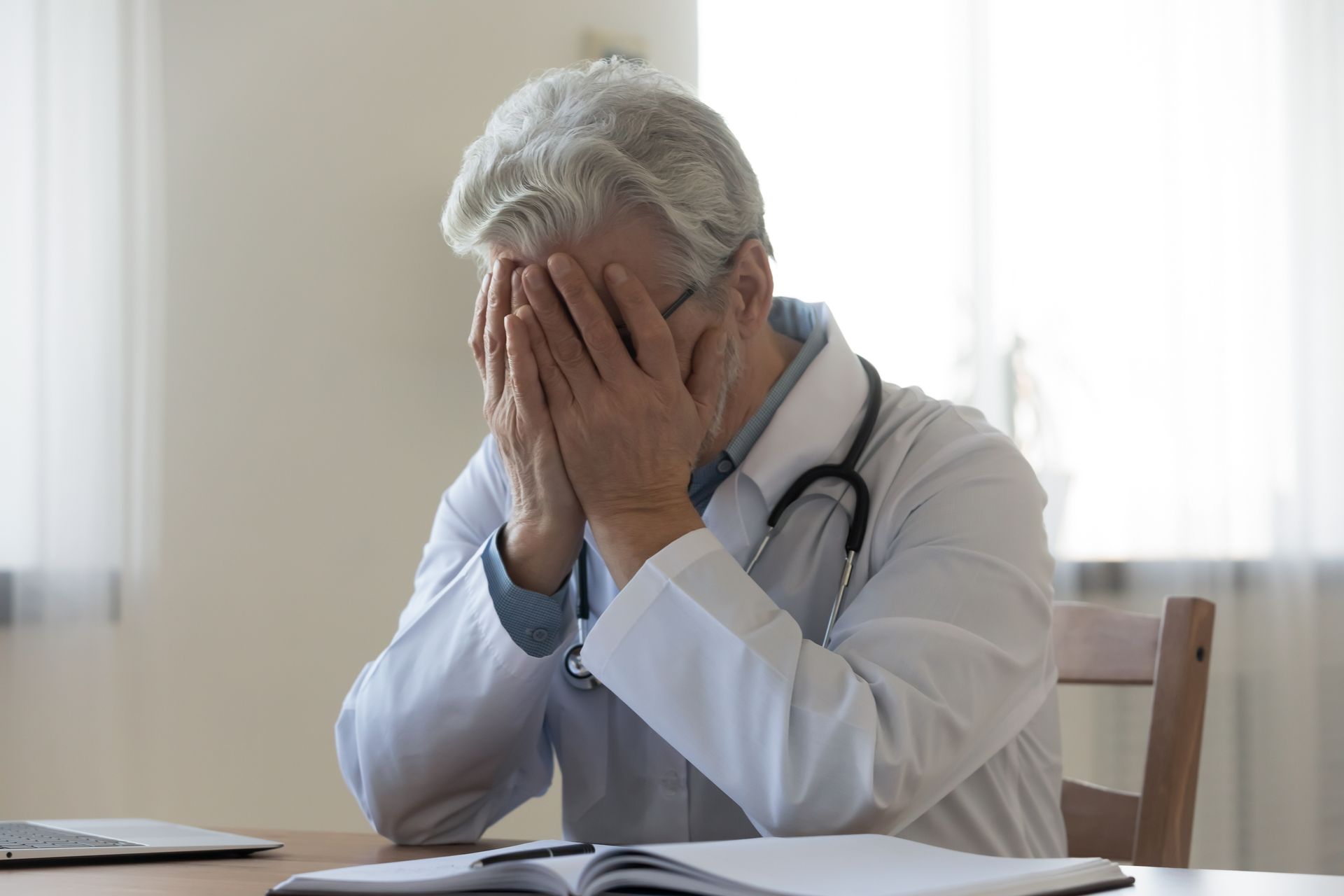 A doctor is sitting at a table with his hands on his face.