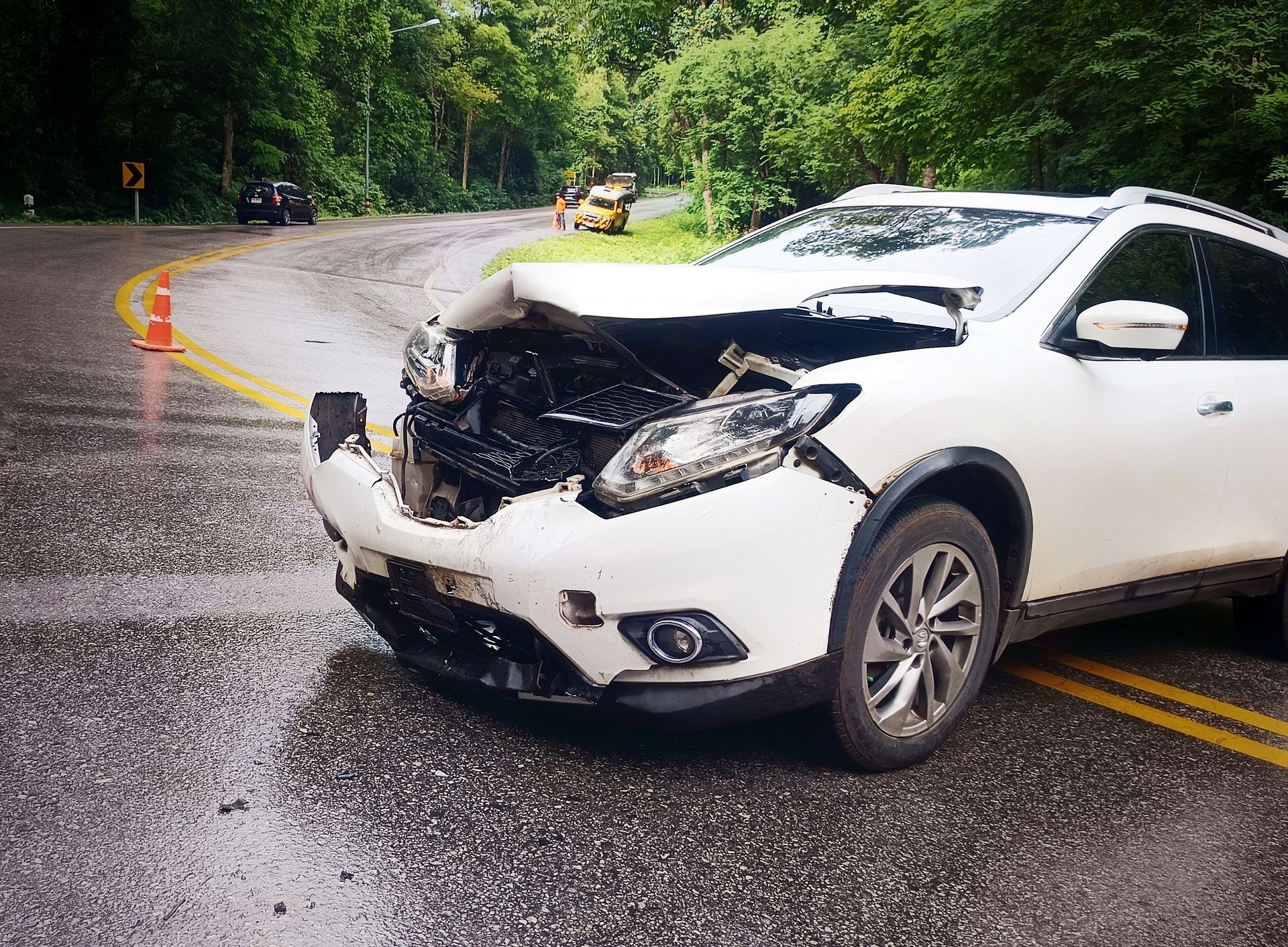 White SUV damaged in a car accident on a wet, winding road. Forest in the background.